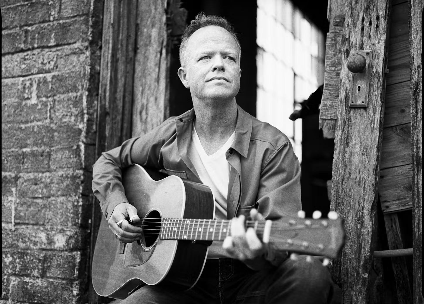 Man playing acoustic guitar outside rustic building, seated in natural light.