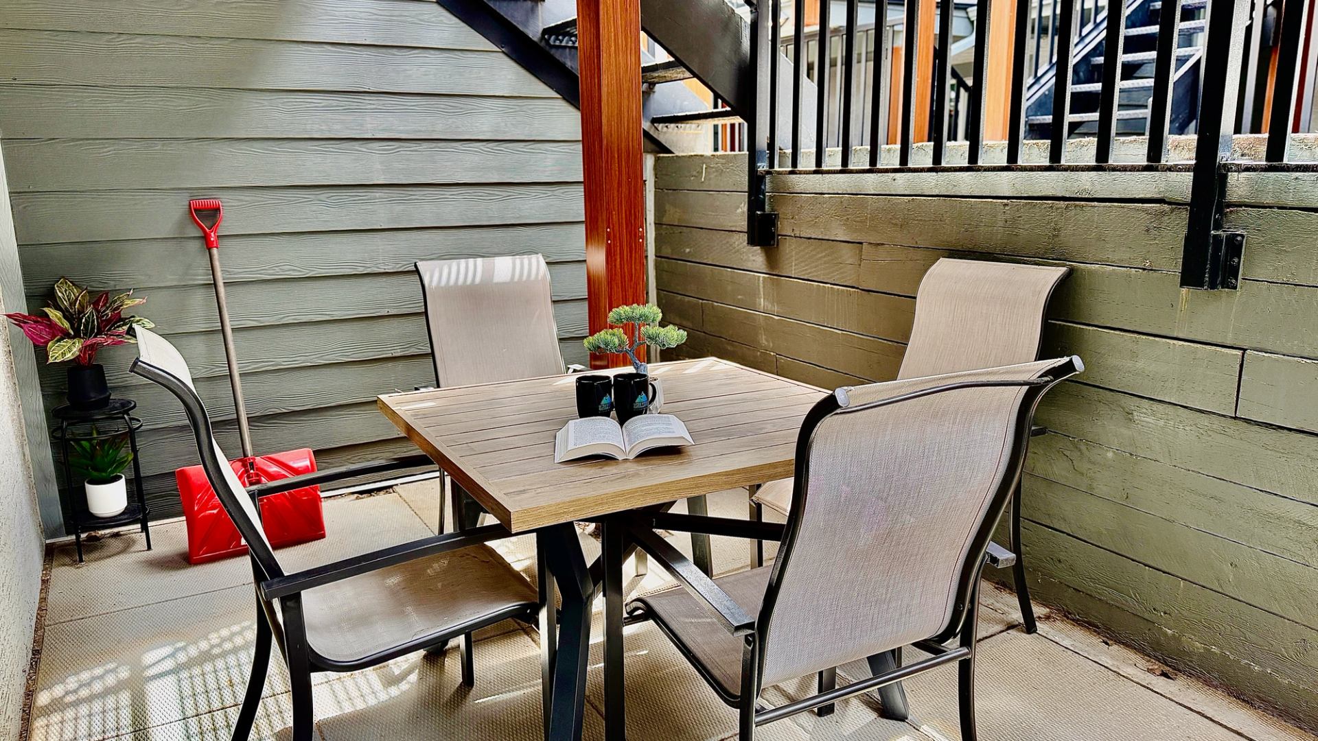 Patio table with four chairs, an open book, mugs and potted plants.