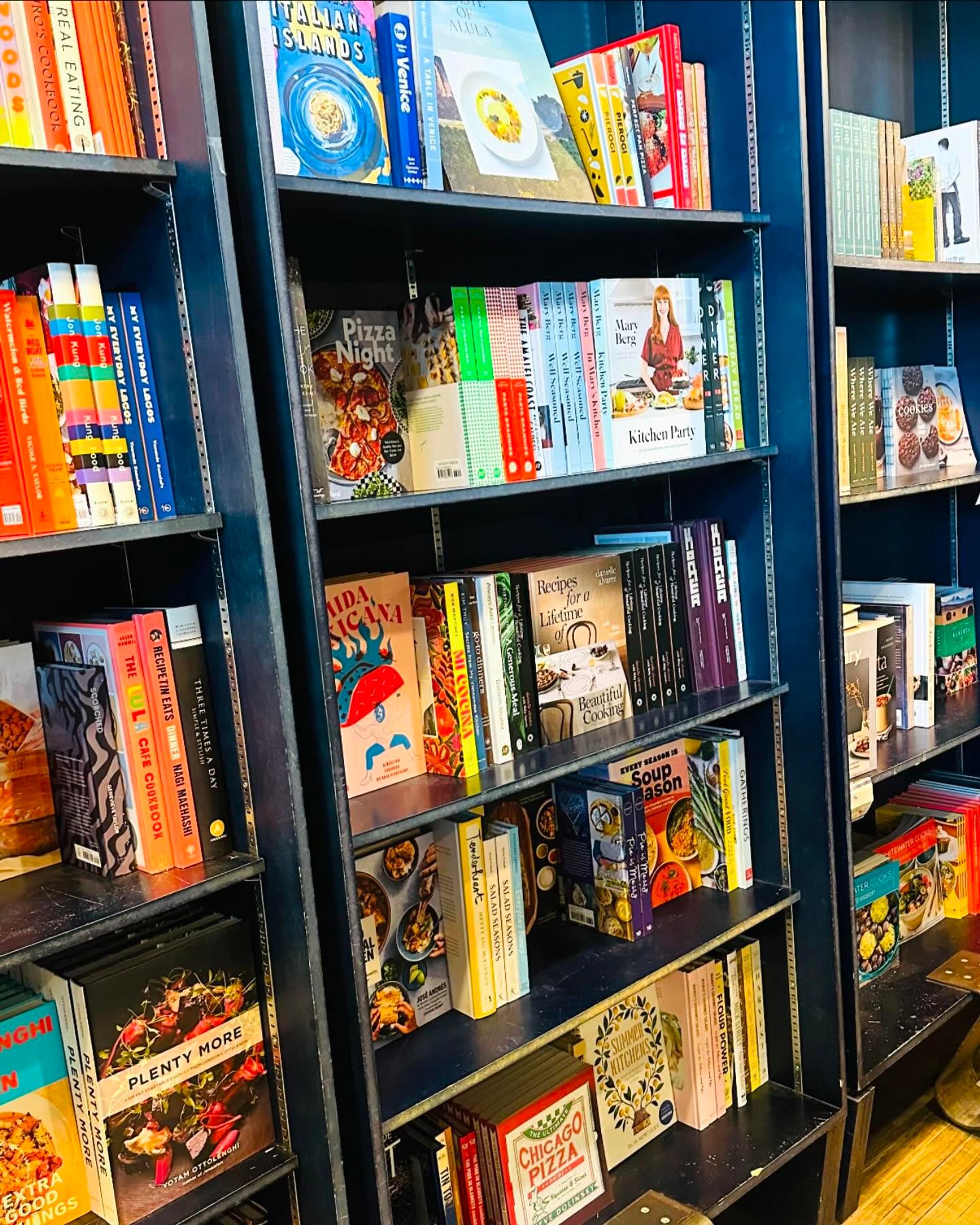 Bookshelves filled with colourful cookbooks and food titles arranged upright and stacked horizontally.