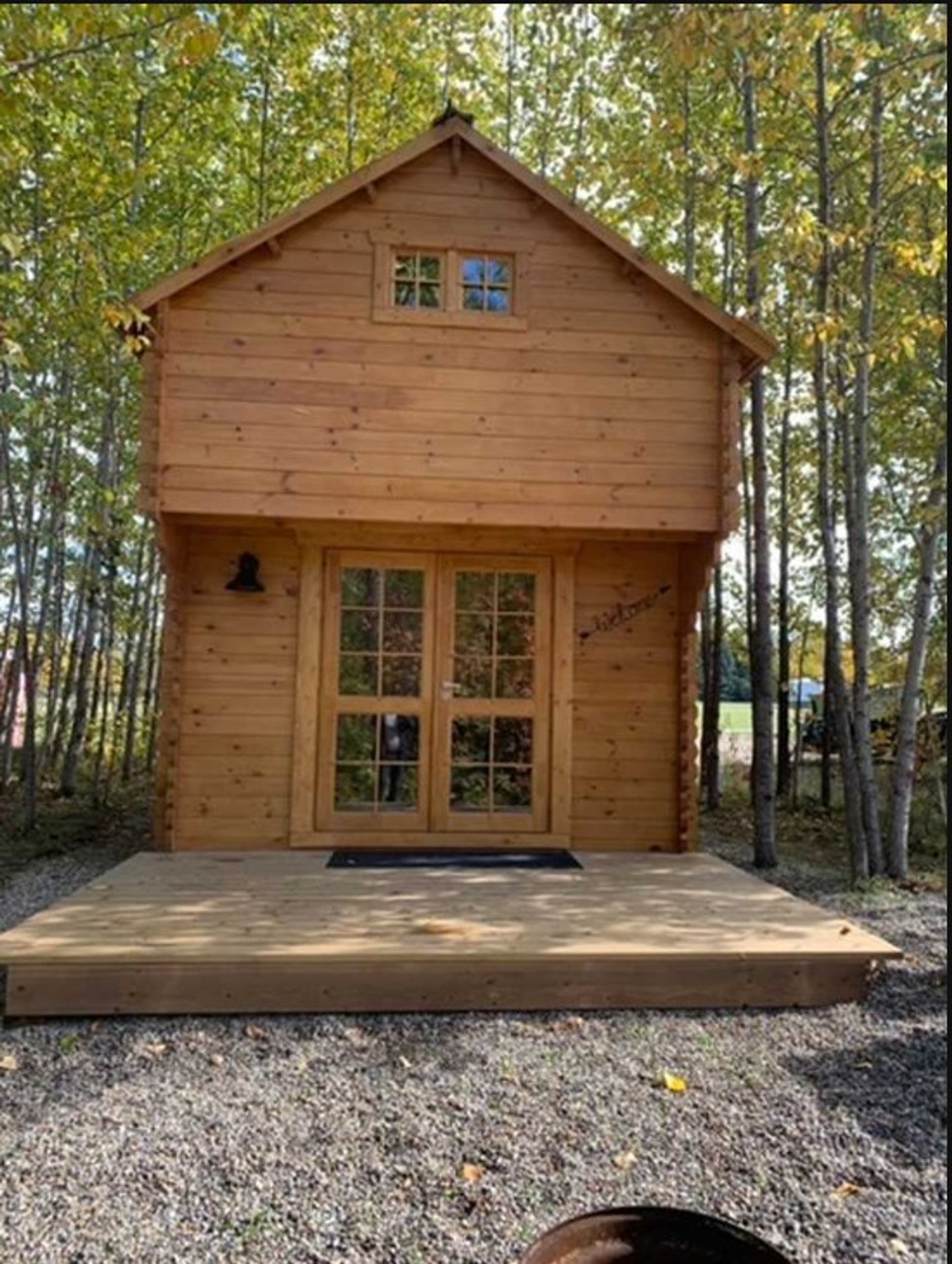 Small wooden cabin with a gabled roof, a front deck, double glass doors, and two small windows, surrounded by autumn trees.