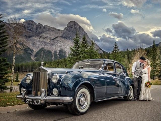 Bride and groom beside classic blue Rolls-Royce in scenic mountain setting.