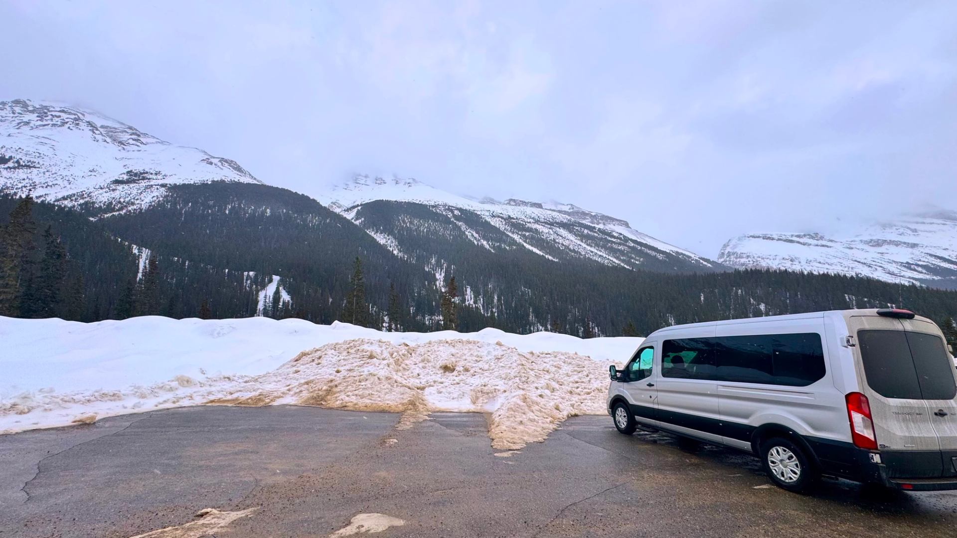 Silver passenger van parked by snowy mountains and evergreen forest under overcast sky.