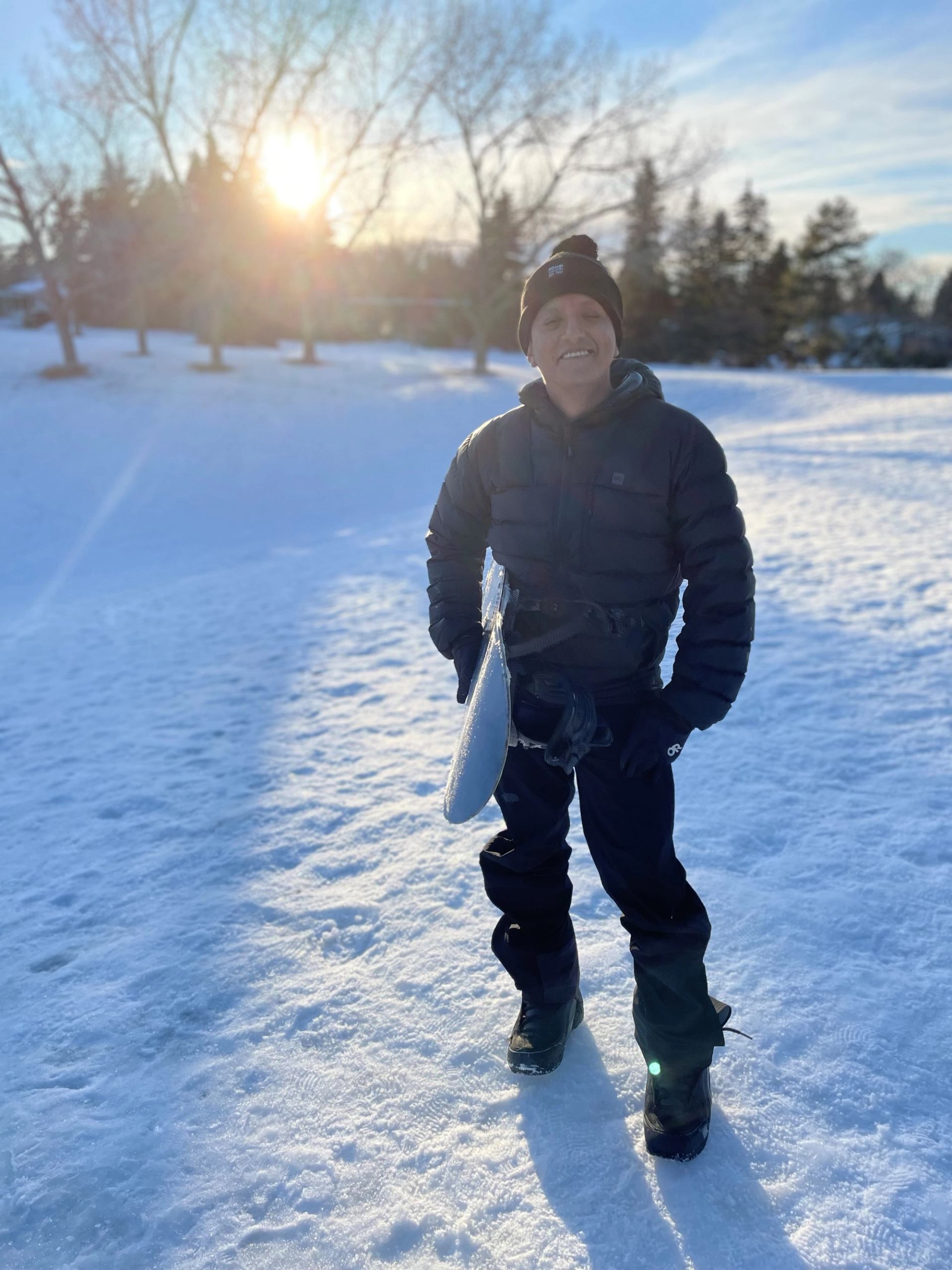 Person in winter gear holding a snowboard on a snowy field, with trees in background.