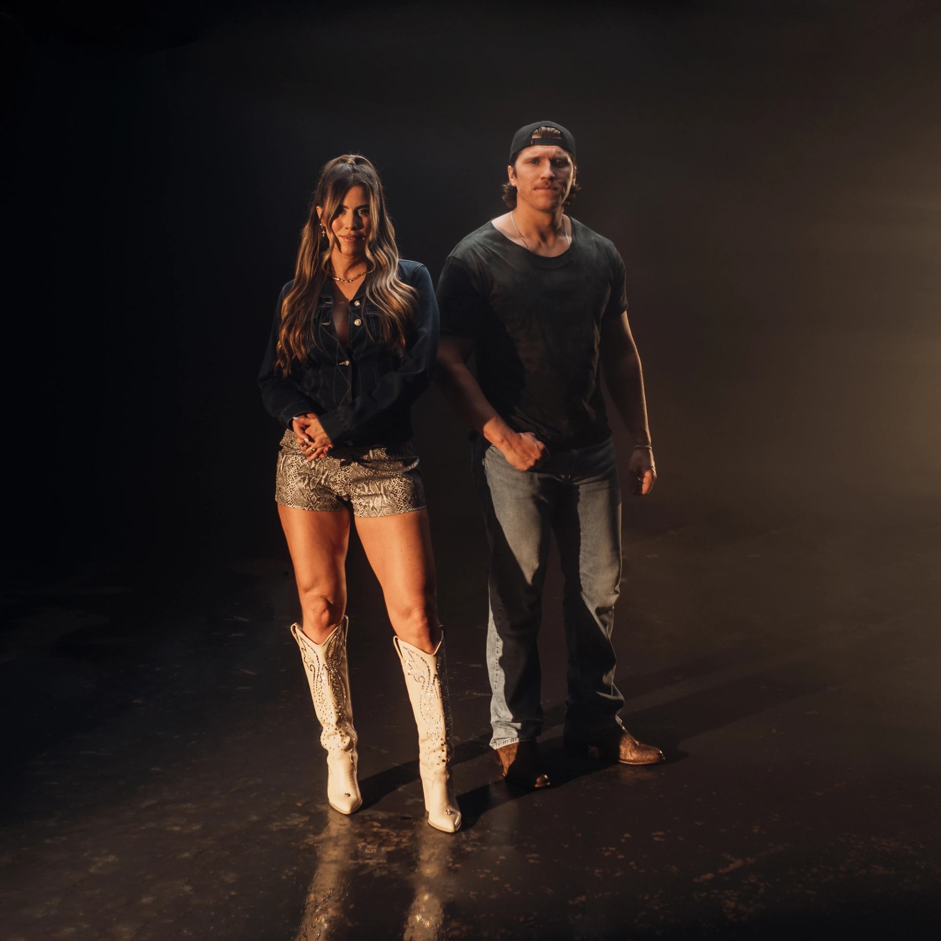 Two people in casual Western clothing stand together against a dark background.