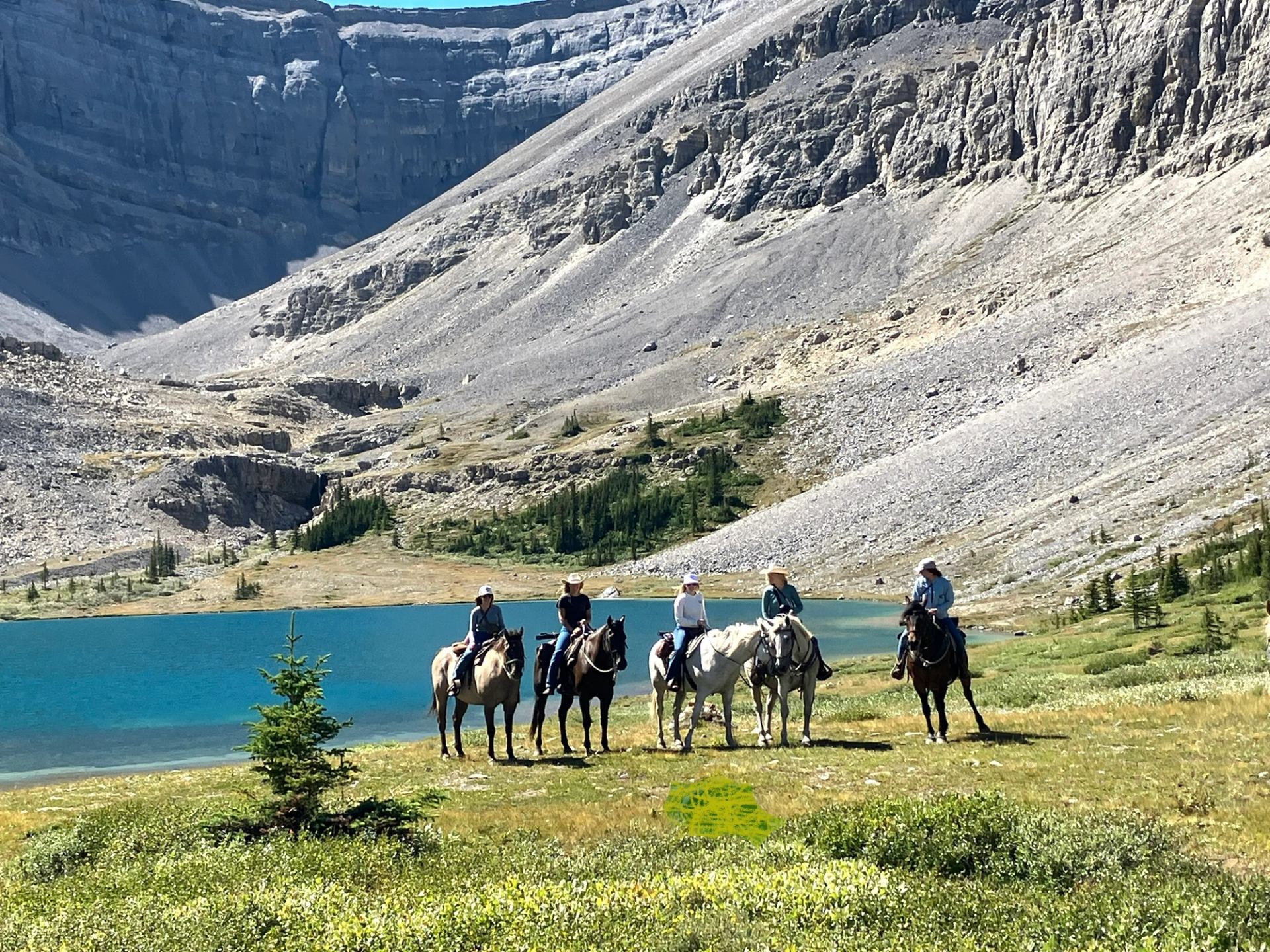 A group of horseback riders pause in a grassy alpine meadow beside a bright blue mountain lake and steep rocky slopes.