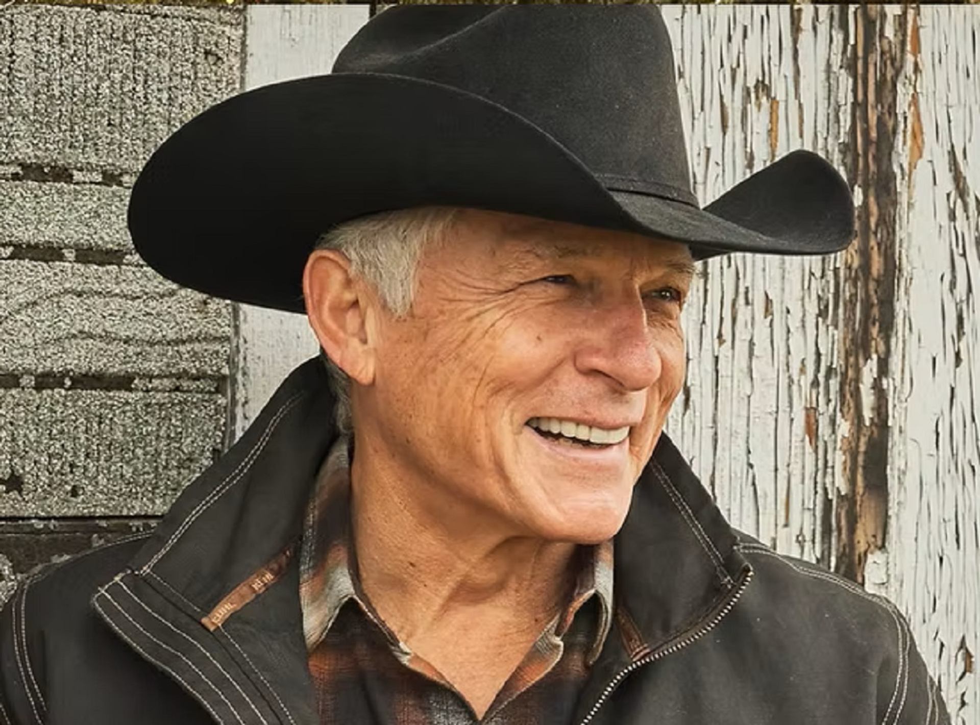Cowboy in black hat and jacket stands before rustic wooden wall.