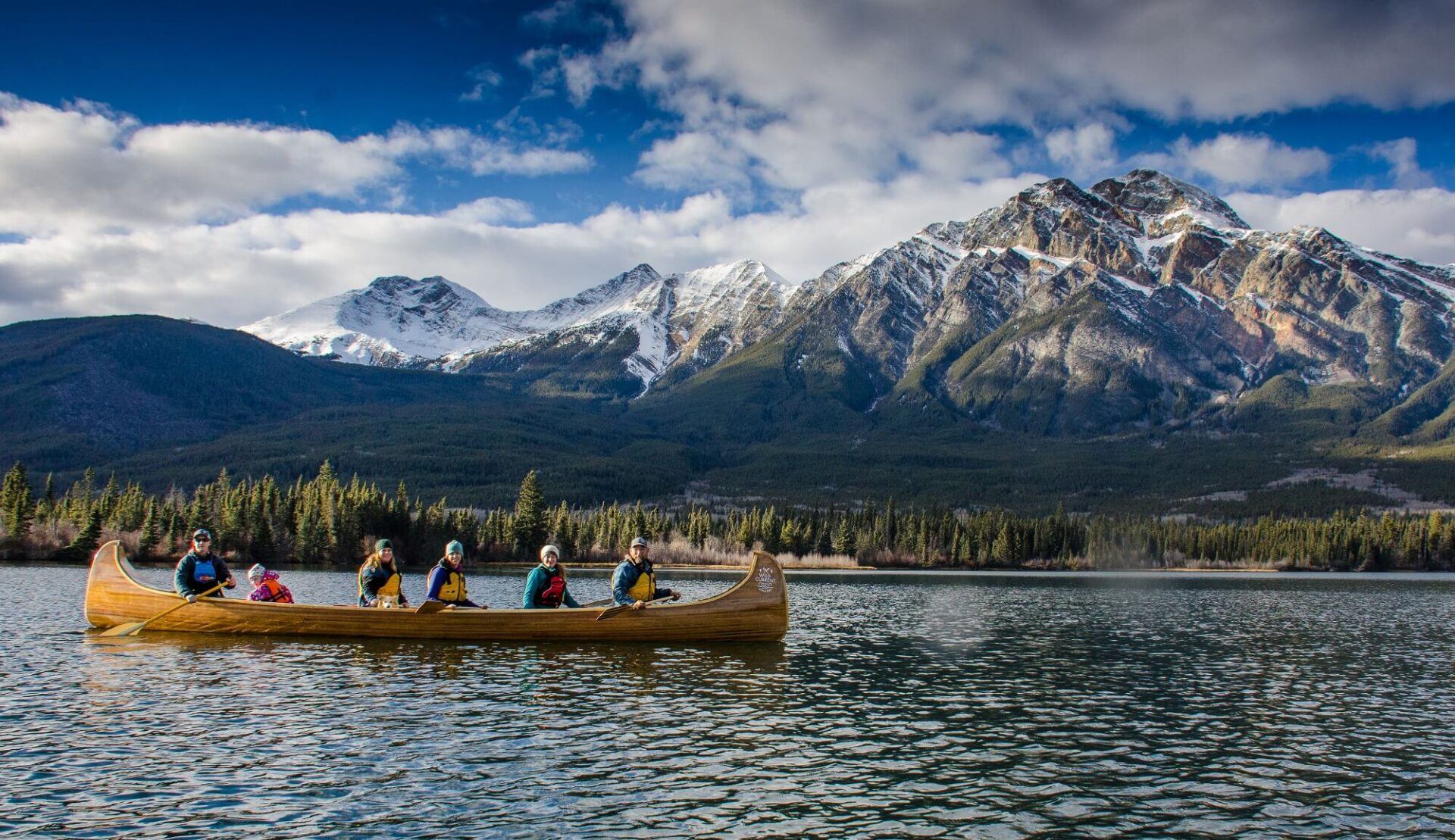 Group paddles a large wooden canoe across a calm mountain lake with snow-dusted peaks behind.