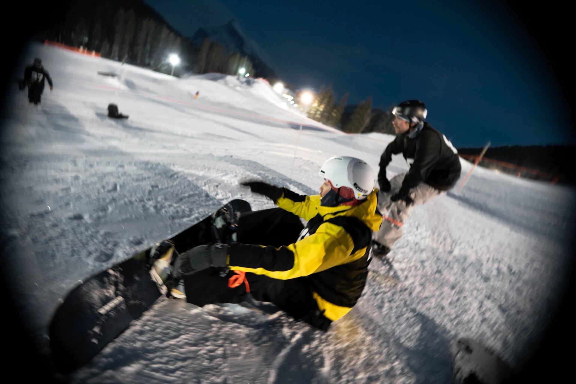 Snowboarder in yellow jacket sitting on the slope during Backyard Rail Jam at night.
