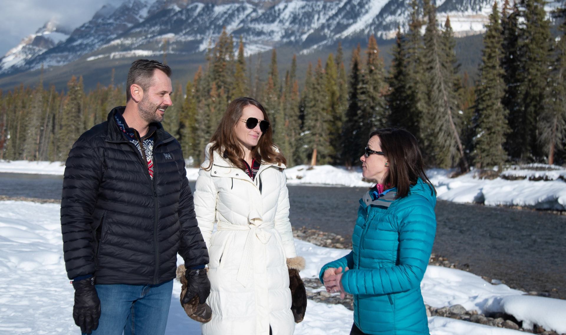 Three people standing near a river with snow-covered banks and towering mountain backdrop