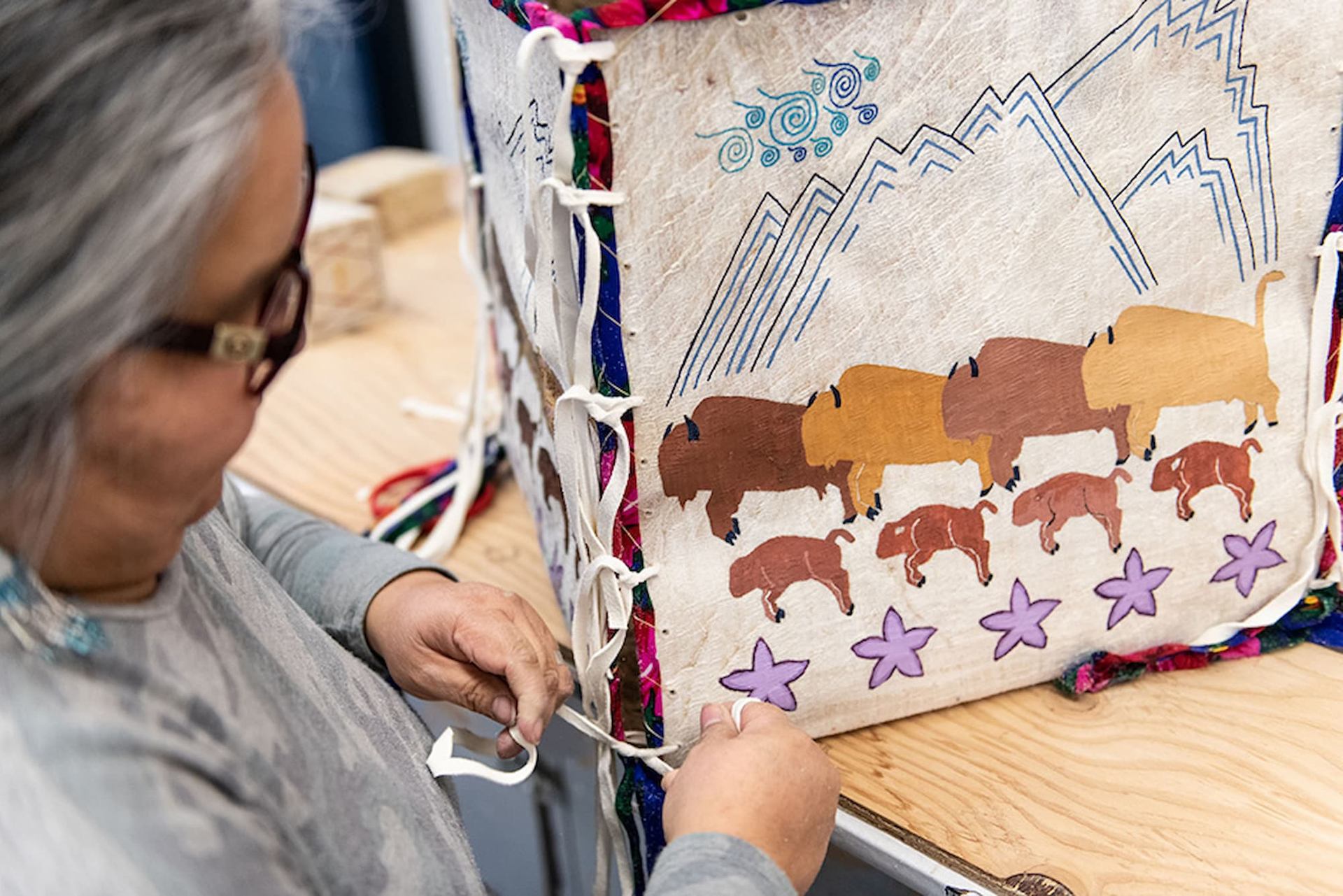 Person laces decorated hide panel with painted bison, mountains and purple flowers.
