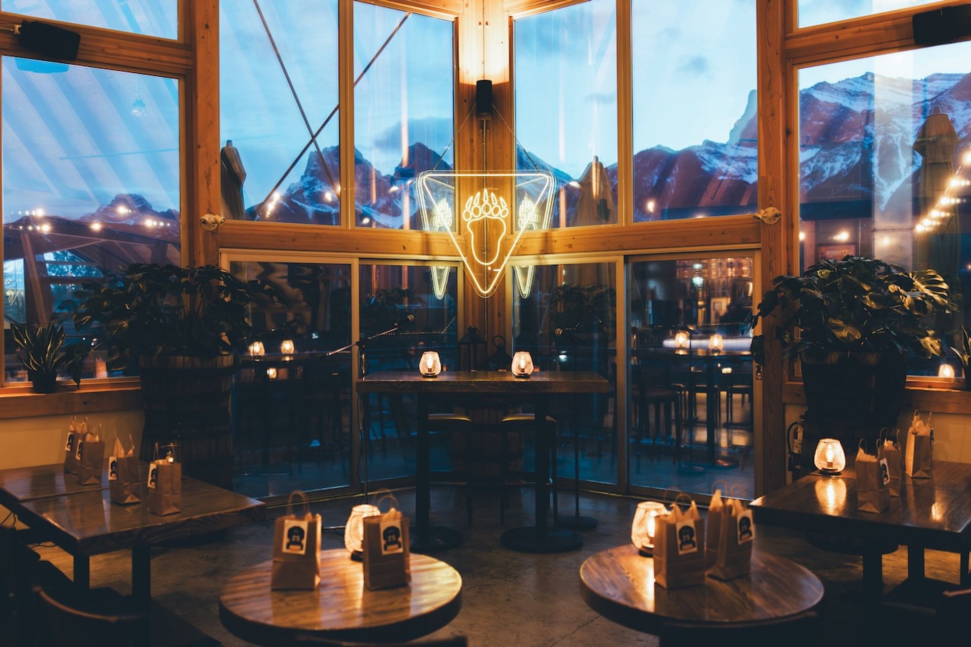 Cozy restaurant interior with large windows overlooking snowy mountains at dusk, featuring an acorn neon sign.
