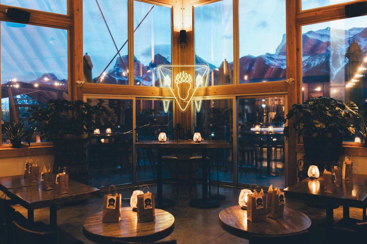 Cozy restaurant interior with large windows overlooking snowy mountains at dusk, featuring an acorn neon sign.