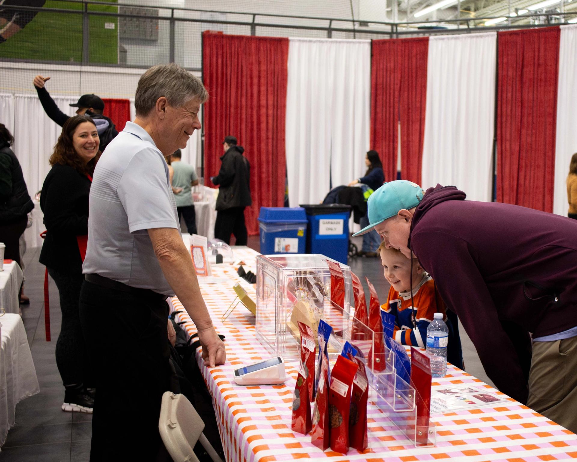 Indoor market table with vendor talking to visitors near clear display boxes and packages.