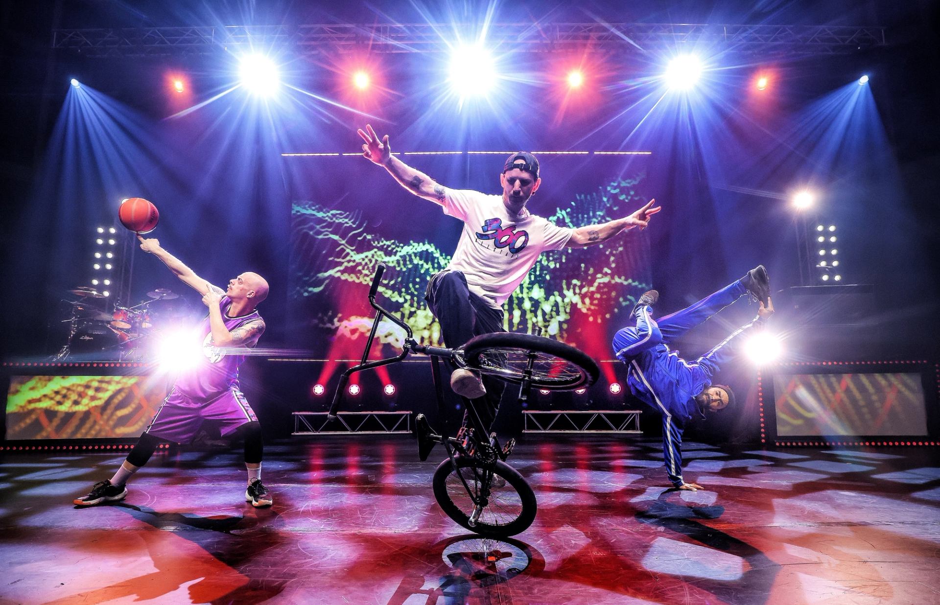 Stage performers do BMX stunt, basketball trick and breakdance under bright concert lights.