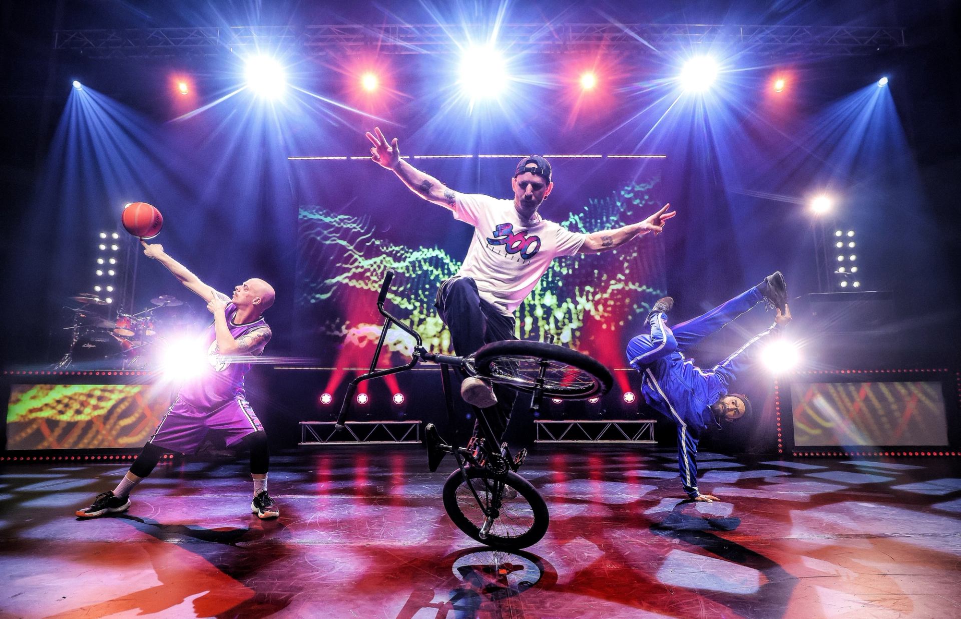 Stage performers do BMX stunt, basketball trick and breakdance under bright concert lights.