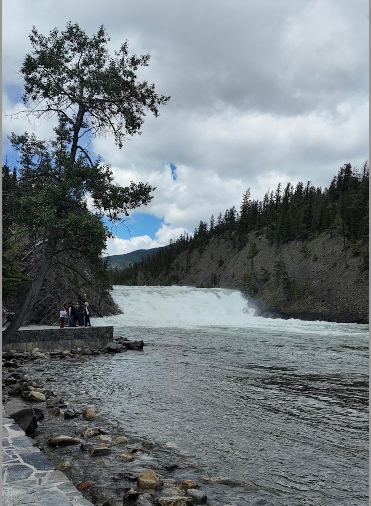 Wide waterfall between forested hills with people standing on stone lookout beside river.