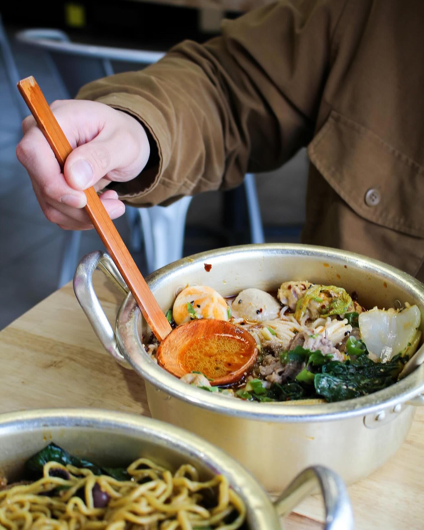 Close-up of ramen bowl with toppings, person holding wooden spoon, second bowl in foreground.