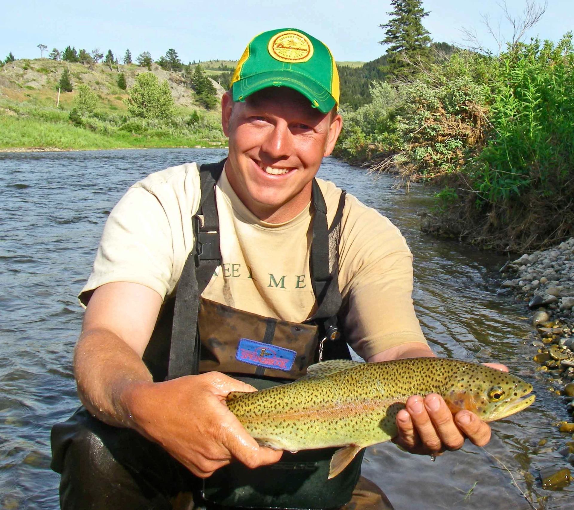 Person in fishing waders holds a trout while standing in a shallow river.