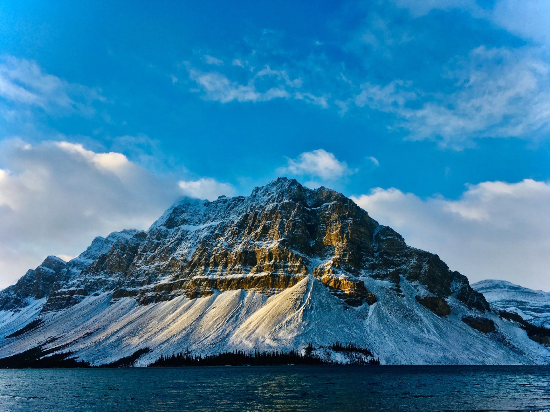 Snow-dusted mountain ridge above dark lake under blue sky with scattered clouds.