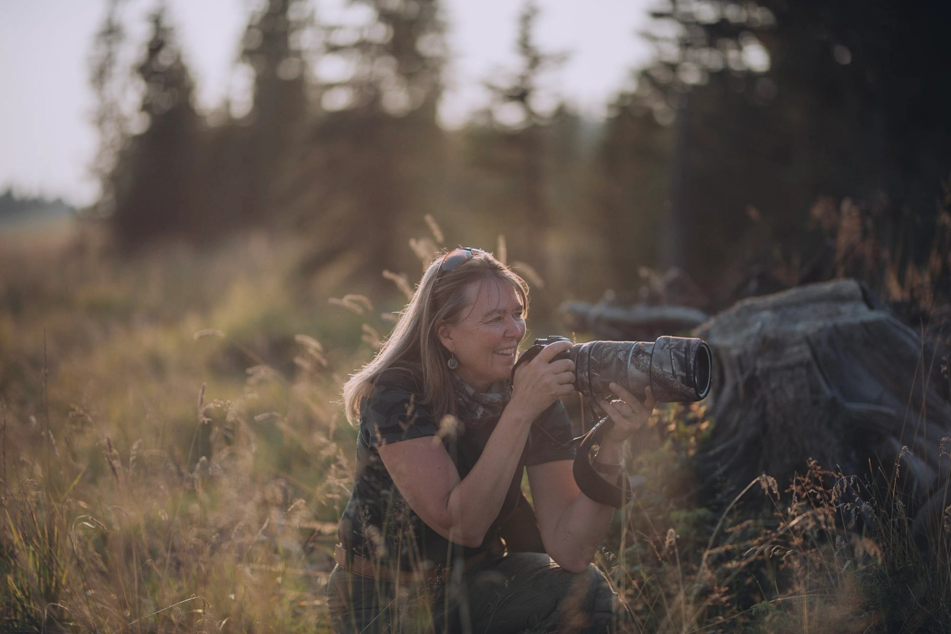 Photographer kneels in tall grass, holding camera with long lens near forest edge