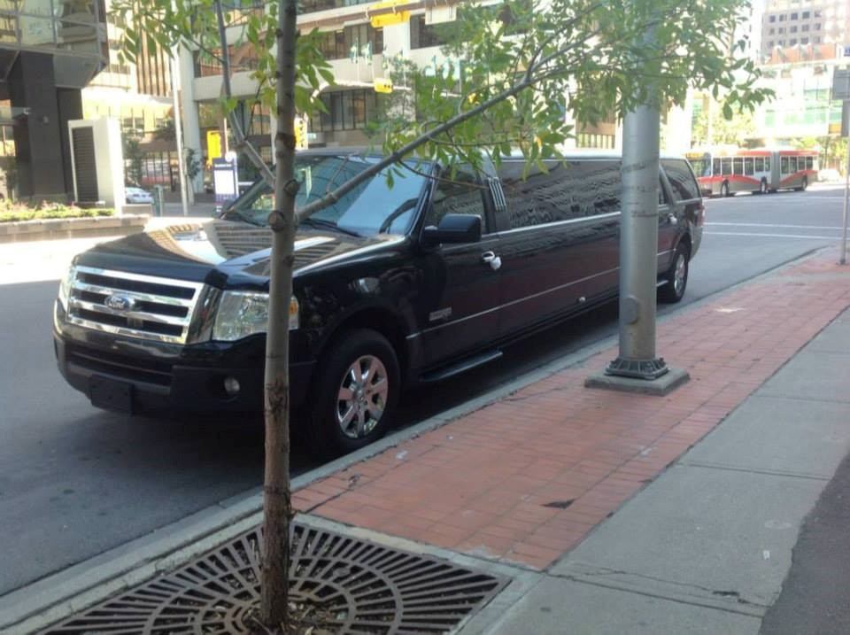 Black stretch limo parked by red-paved sidewalk in city, tree and light pole in foreground.