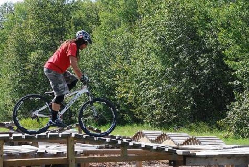 Mountain biker rides over wooden ramp feature with trees in background.