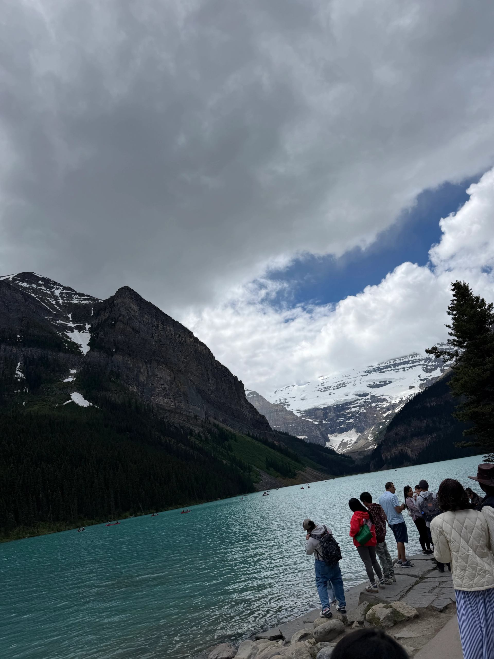 Tourists stand along turquoise mountain lake shore under cloudy sky with snow-capped peaks.