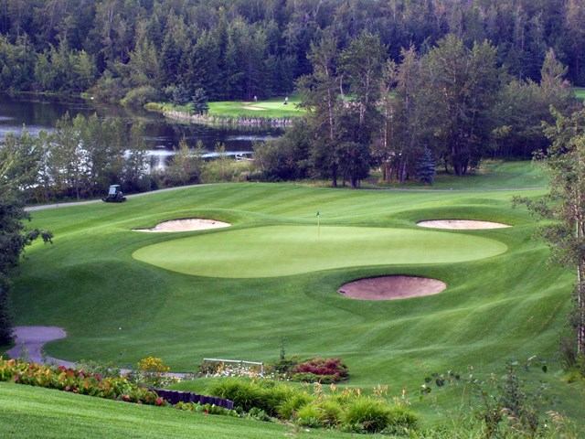 Golf green with flagstick, sand bunkers, trees and pond in background.