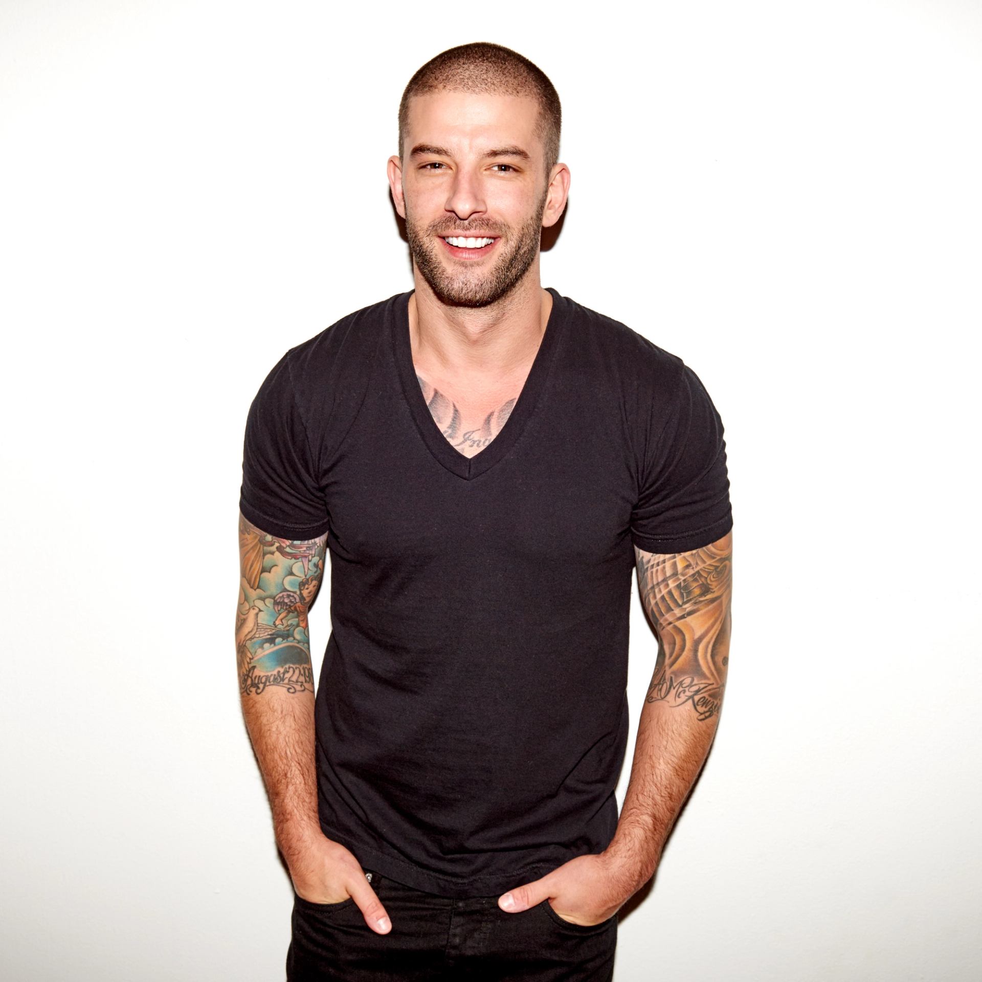 Tattooed person in black V-neck T-shirt stands against plain white background.