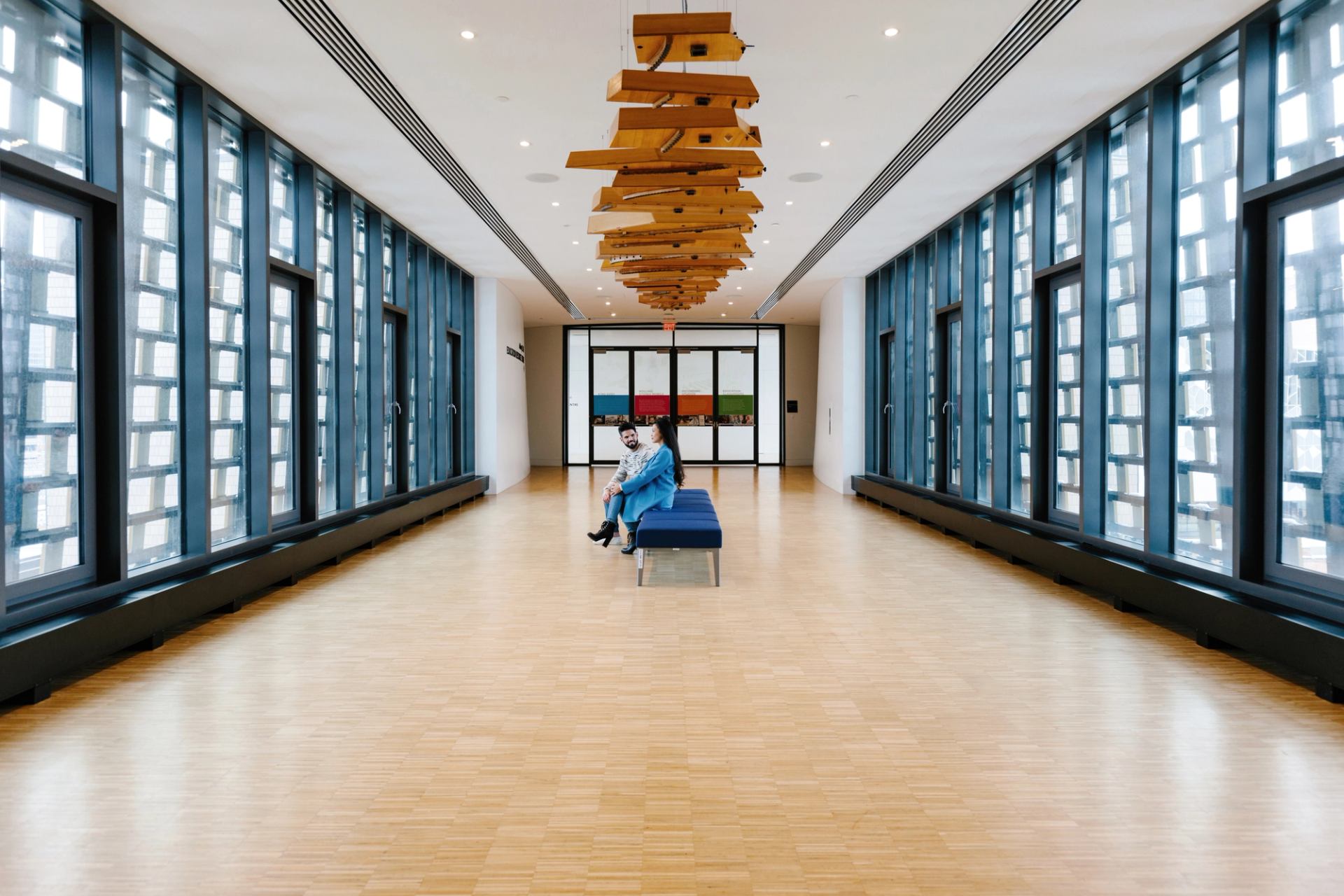 Wide hallway with large windows, wooden ceiling sculpture and two people sitting on bench.