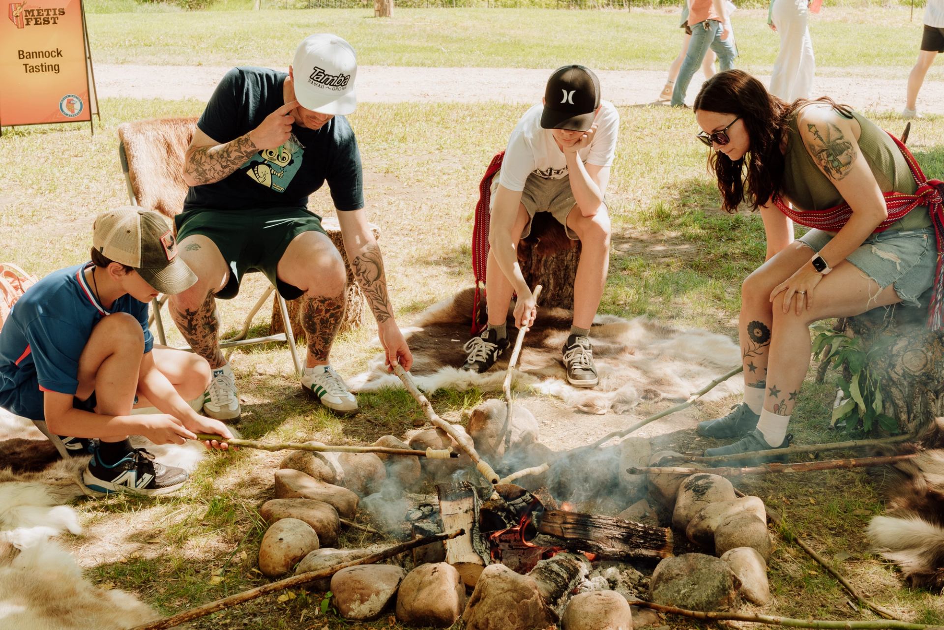 People roast bannock on sticks over a smoky campfire at an outdoor event.