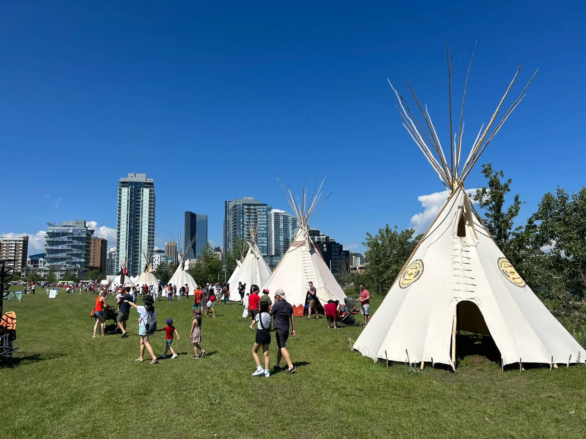 Tipis on a grassy field with people walking, modern city skyline in background.
