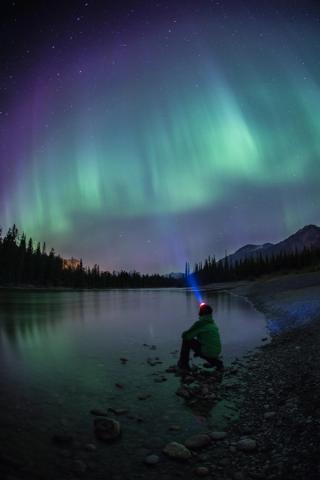 Person with a headlamp sitting near a lake watching the northern lights in the Jasper National Park Dark Sky Preserve.