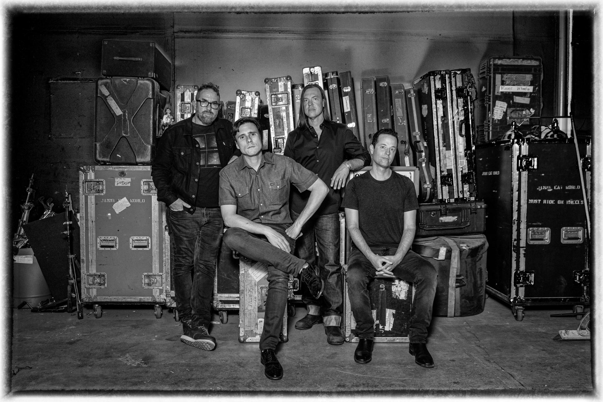 Black and white photo of four men, a band, posing among road cases.