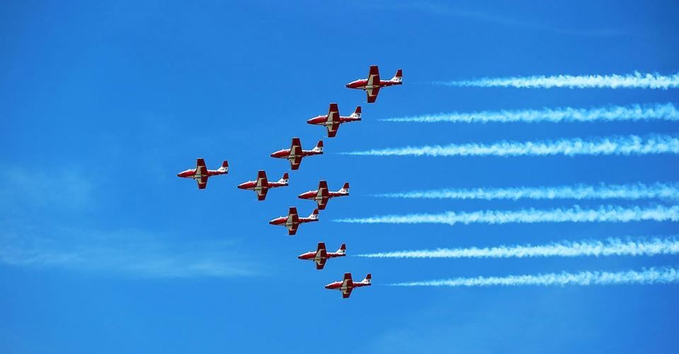 Red and white aerobatic jets fly in formation, leaving parallel white smoke trails.