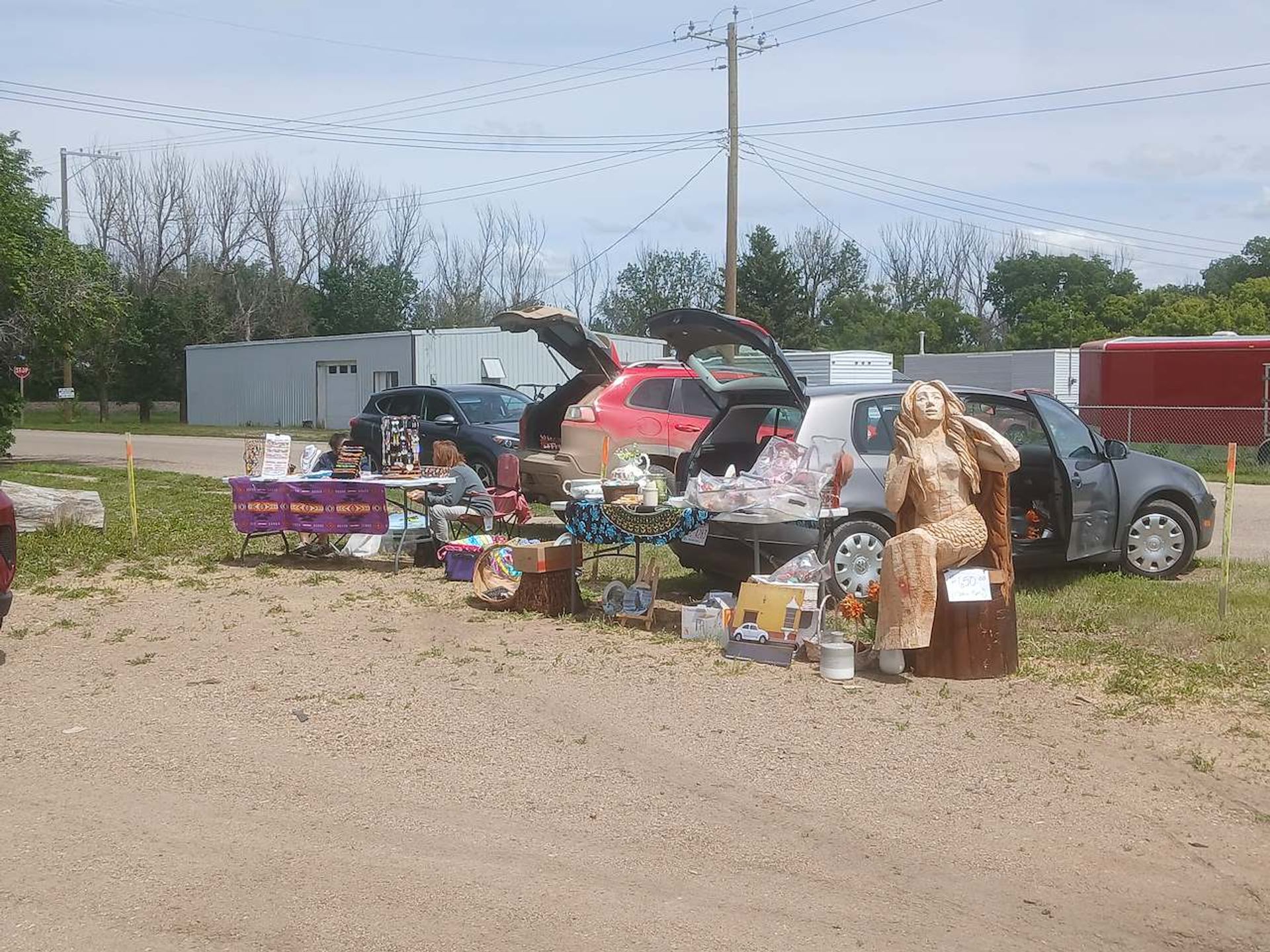 Outdoor roadside market with car trunks open, tables of crafts, and large wooden statue.