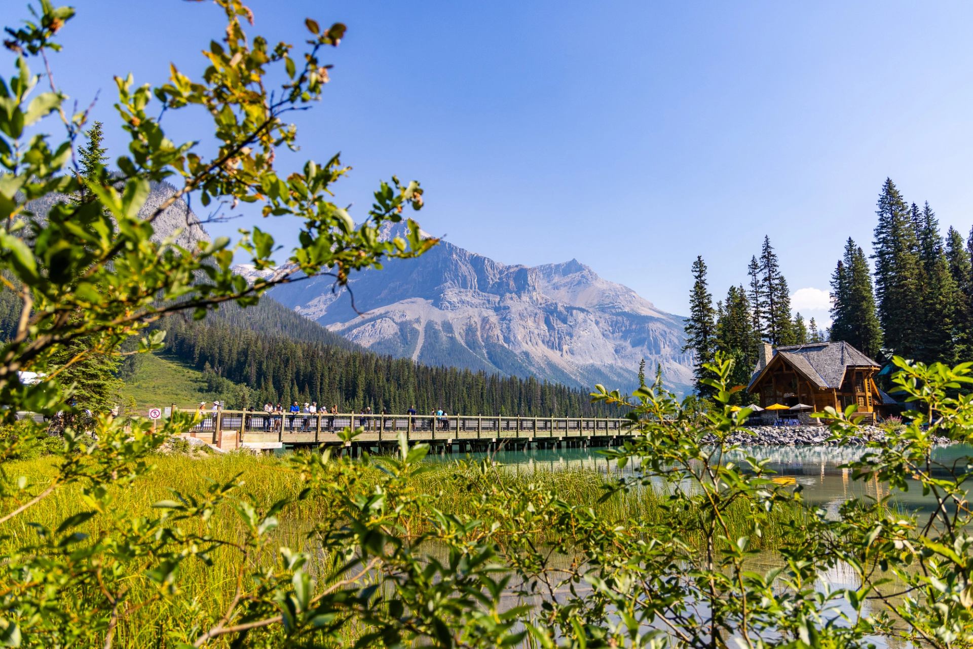 Wooden lodge and bridge over calm lake, framed by shrubs and distant mountains.