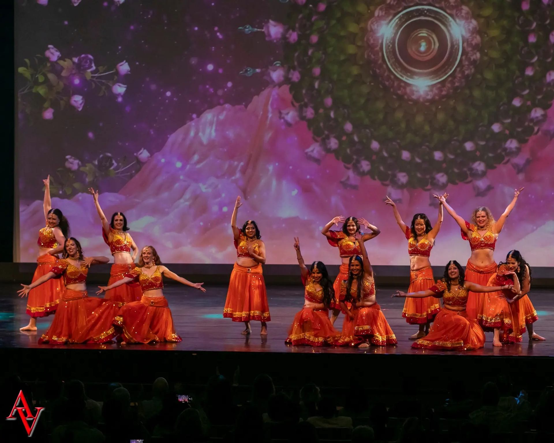 A group of dancers wearing coordinated orange and gold costumes posing on stage, with some standing and others kneeling, in front of a colorful background featuring floral and cosmic imagery.