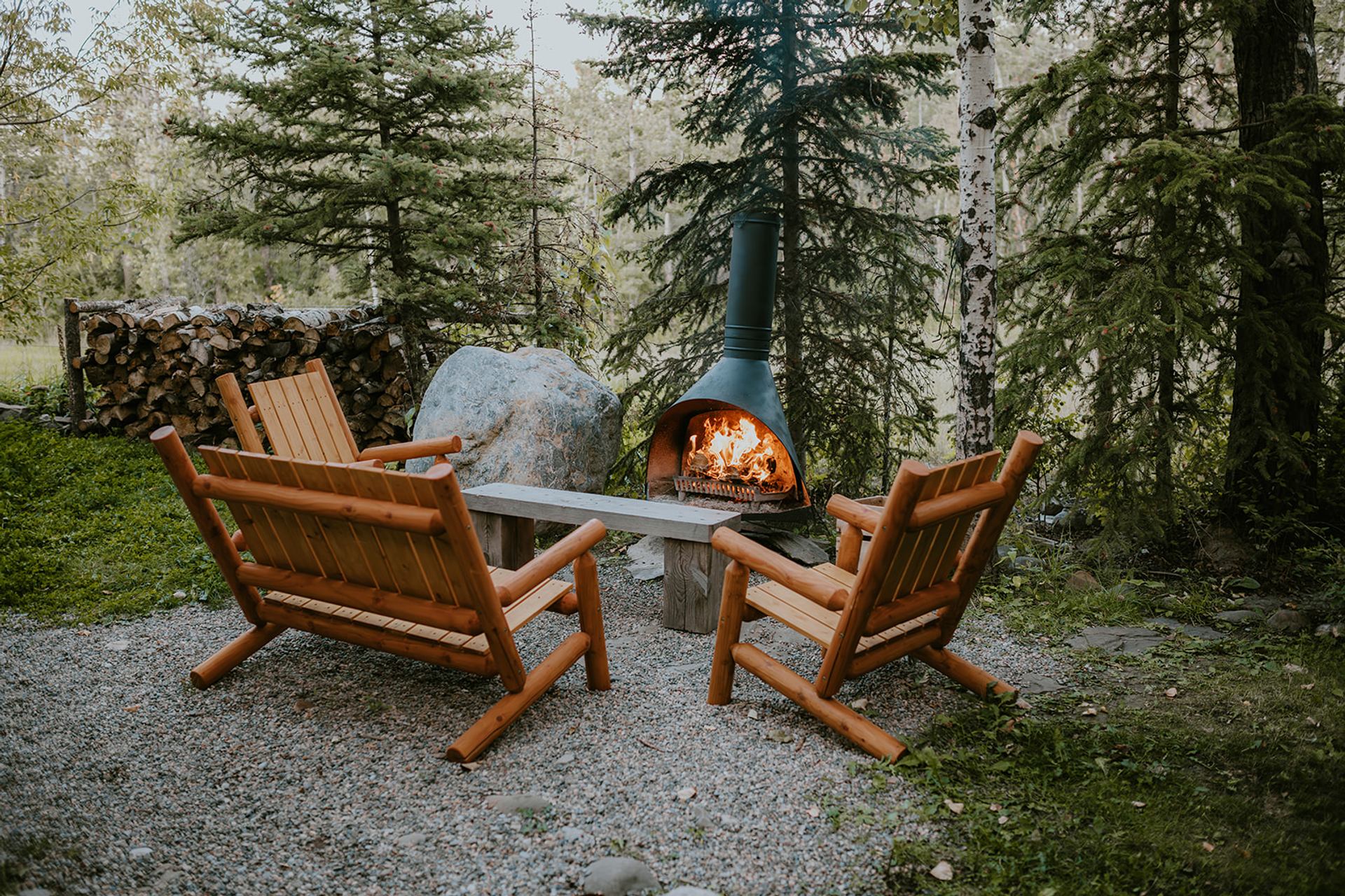 Wooden chairs surround a burning outdoor fireplace in a forest clearing.