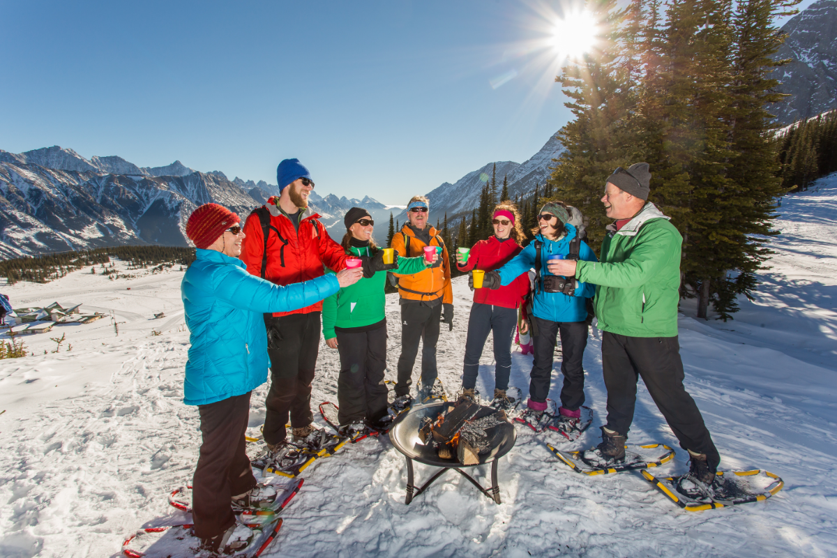 Snowshoers stand around a small fire pit, raising cups in snowy mountain landscape..