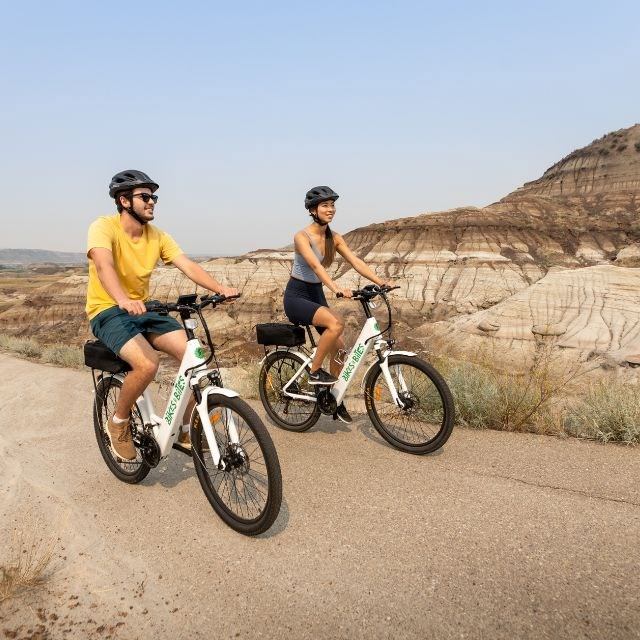 Two people ride electric bikes on a paved trail through scenic badlands with layered rock formations.