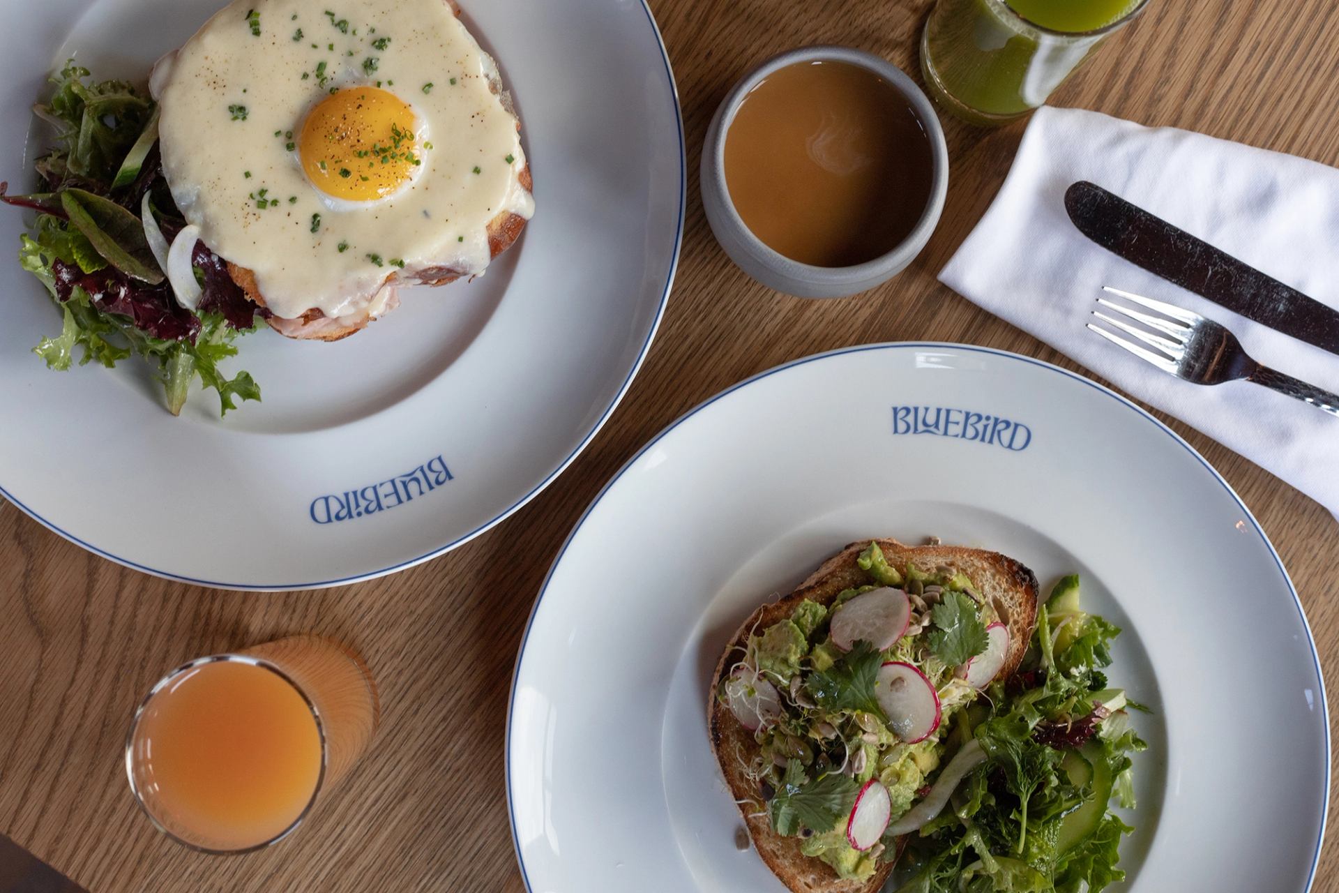 Overhead shot of breakfast with Croque Madame, avocado toast, side salads, juices, and coffee on a wooden table.