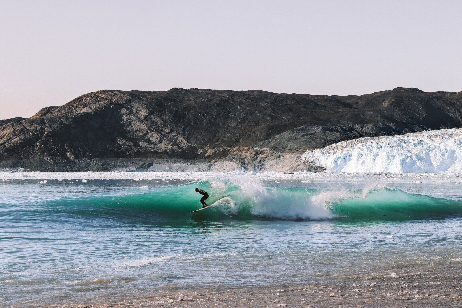 Surfer rides wave with mountain coastline in background, near the shore.