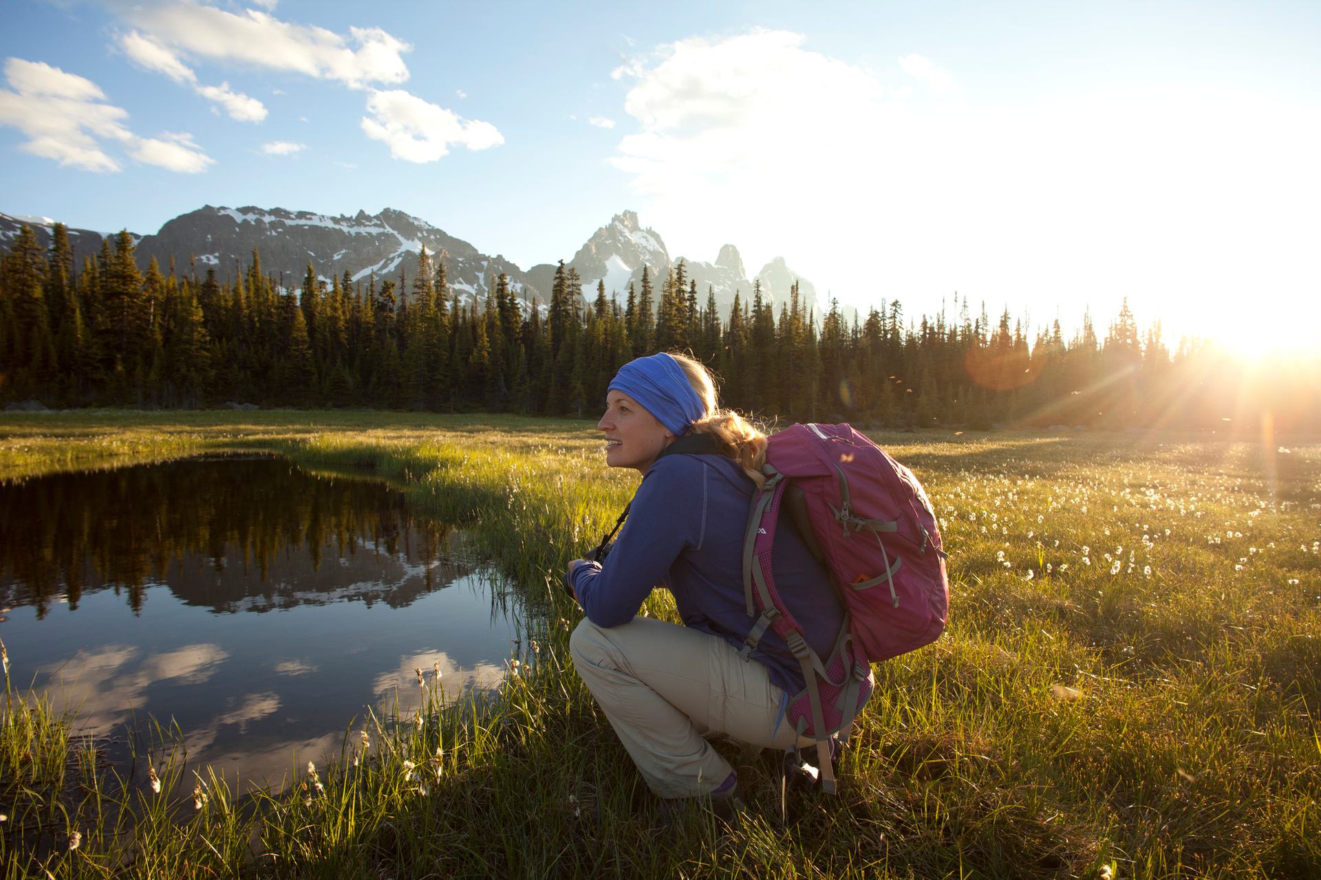 Hiker with a backpack crouches near a reflective pond with mountain peaks and pine forest behind.