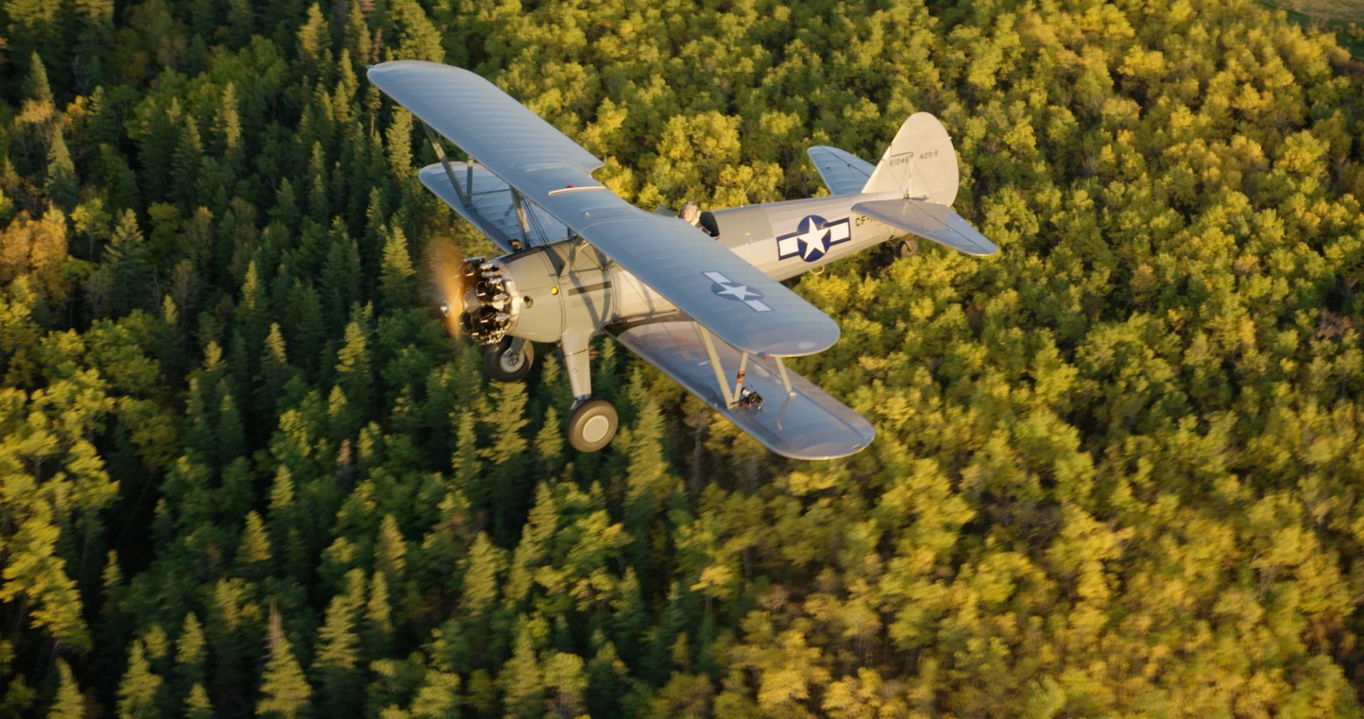 Vintage biplane with military markings flying above dense green forest canopy.