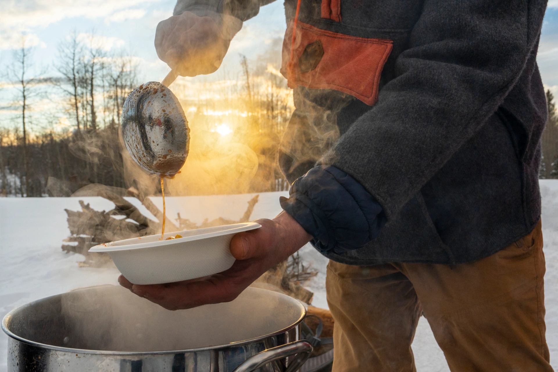 Steaming soup being ladled into a bowl outdoors with snow and sunset in the background