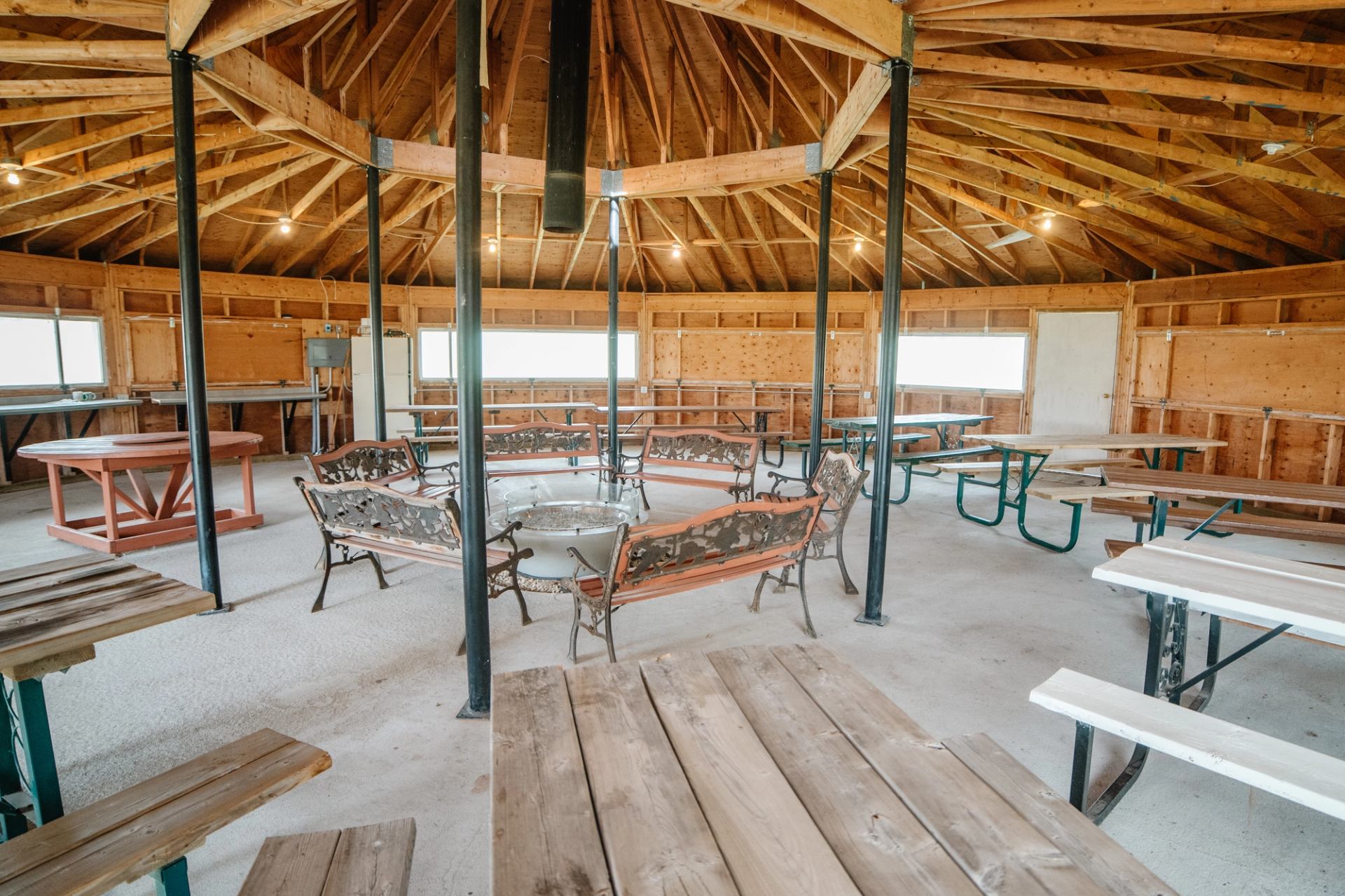 Wooden pavilion interior with picnic tables and benches arranged around a central firepit.