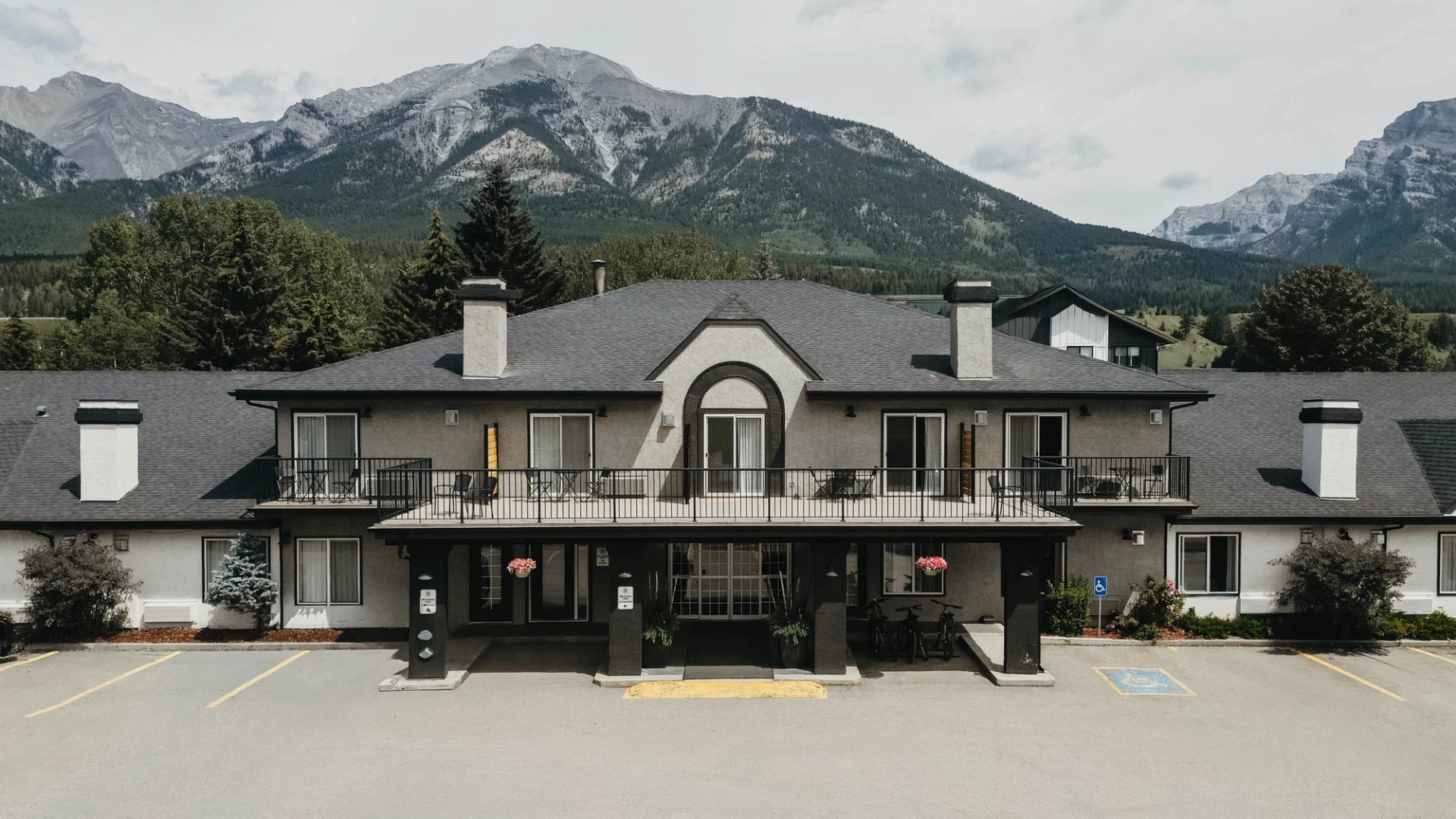 Two-storey lodge-style building with balconies, parking lot and mountain range in background.