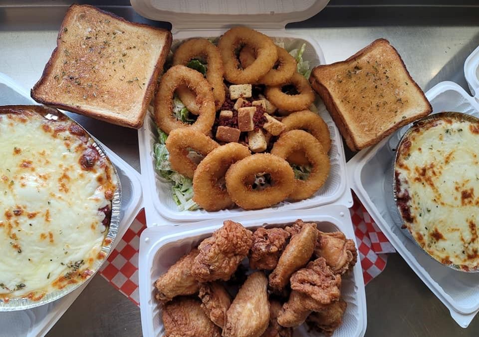 Takeout containers with lasagna, fried chicken, onion rings, salad, and garlic bread.