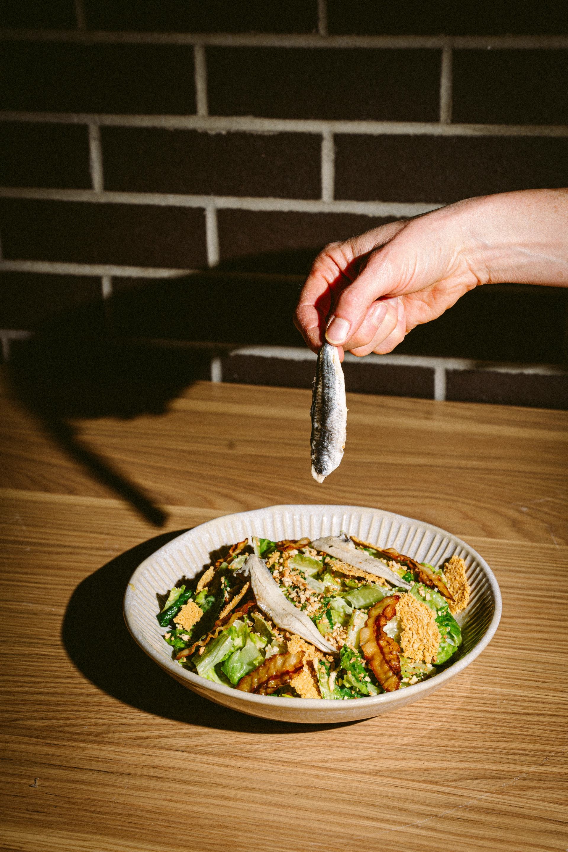 A hand holds a small fish above a salad of greens, crispy toppings, and fish fillets on a wooden table.